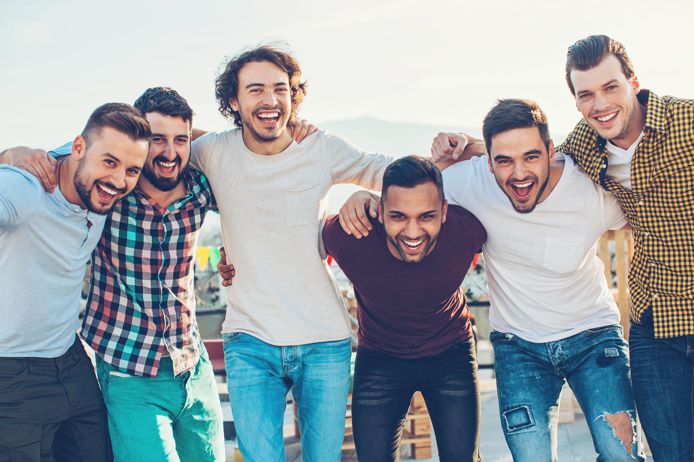 Six men standing together outdoors with their arms around one another, smiling.