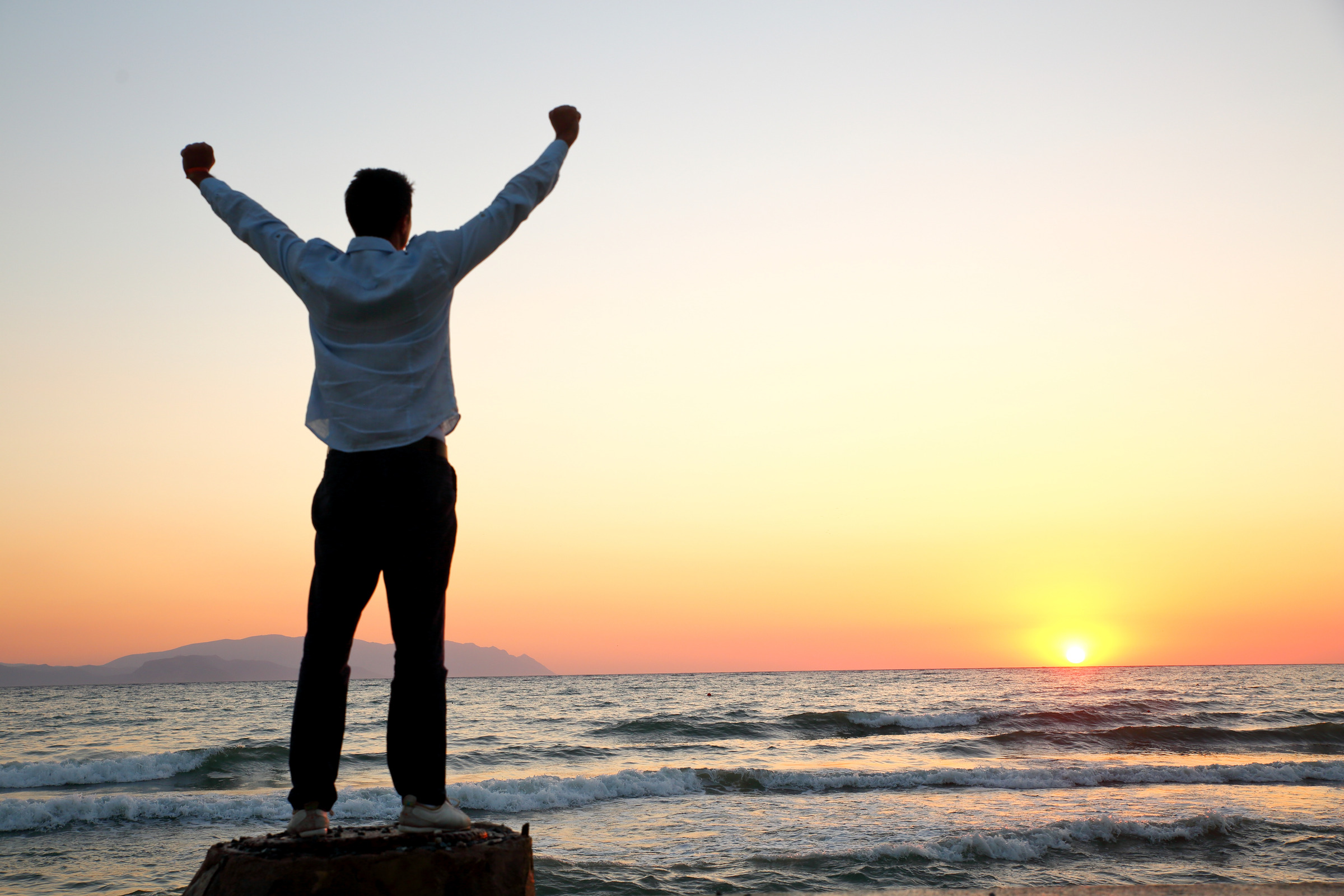 A man standing by the ocean at sunset with his arms raised.