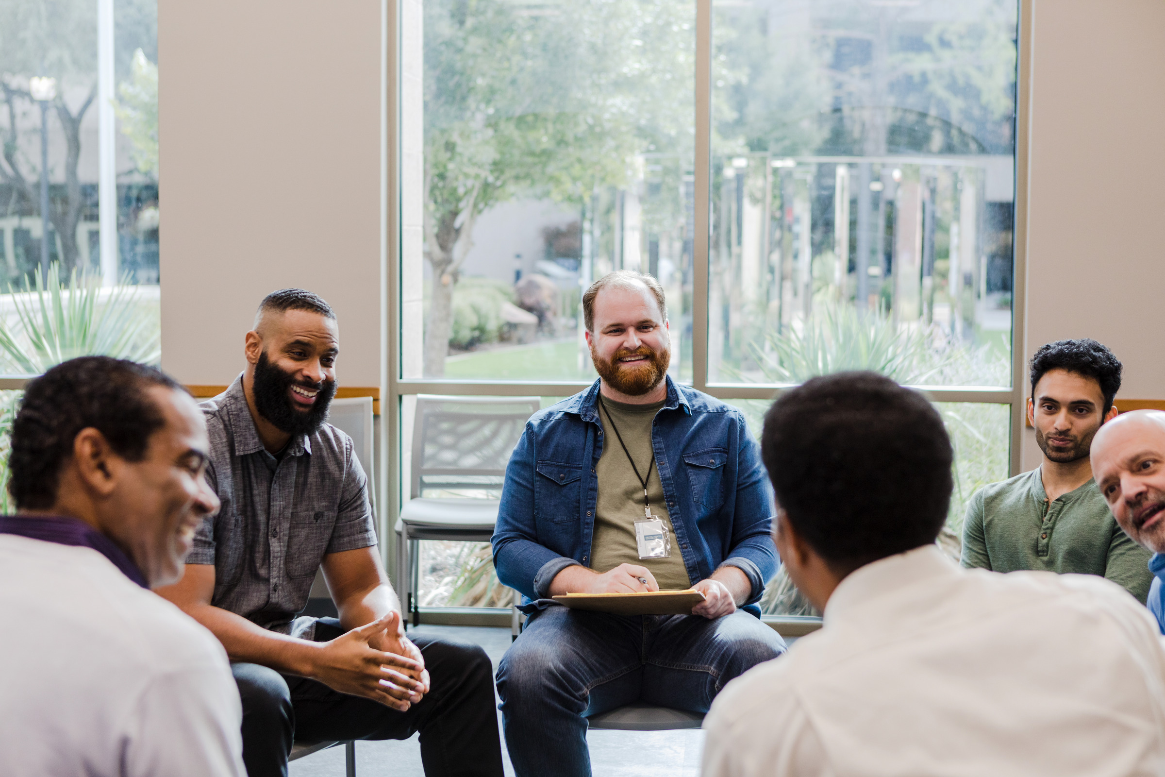 A group of men sitting in a circle in a bright room during a meeting.